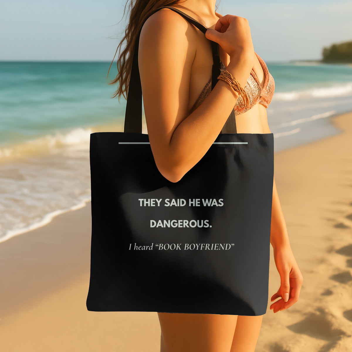 A woman on a sandy beach carries a black tote bag with white text that reads, 'THEY SAID HE WAS DANGEROUS. I heard "BOOK BOYFRIEND"'.