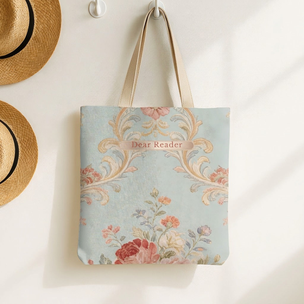 A canvas tote bag with a light blue background, featuring a vintage floral and scroll pattern in soft pink, peach, and beige tones. The bag has beige shoulder straps and a small central label that reads 'Dear Reader'. It is displayed on a hook next to some straw hats.