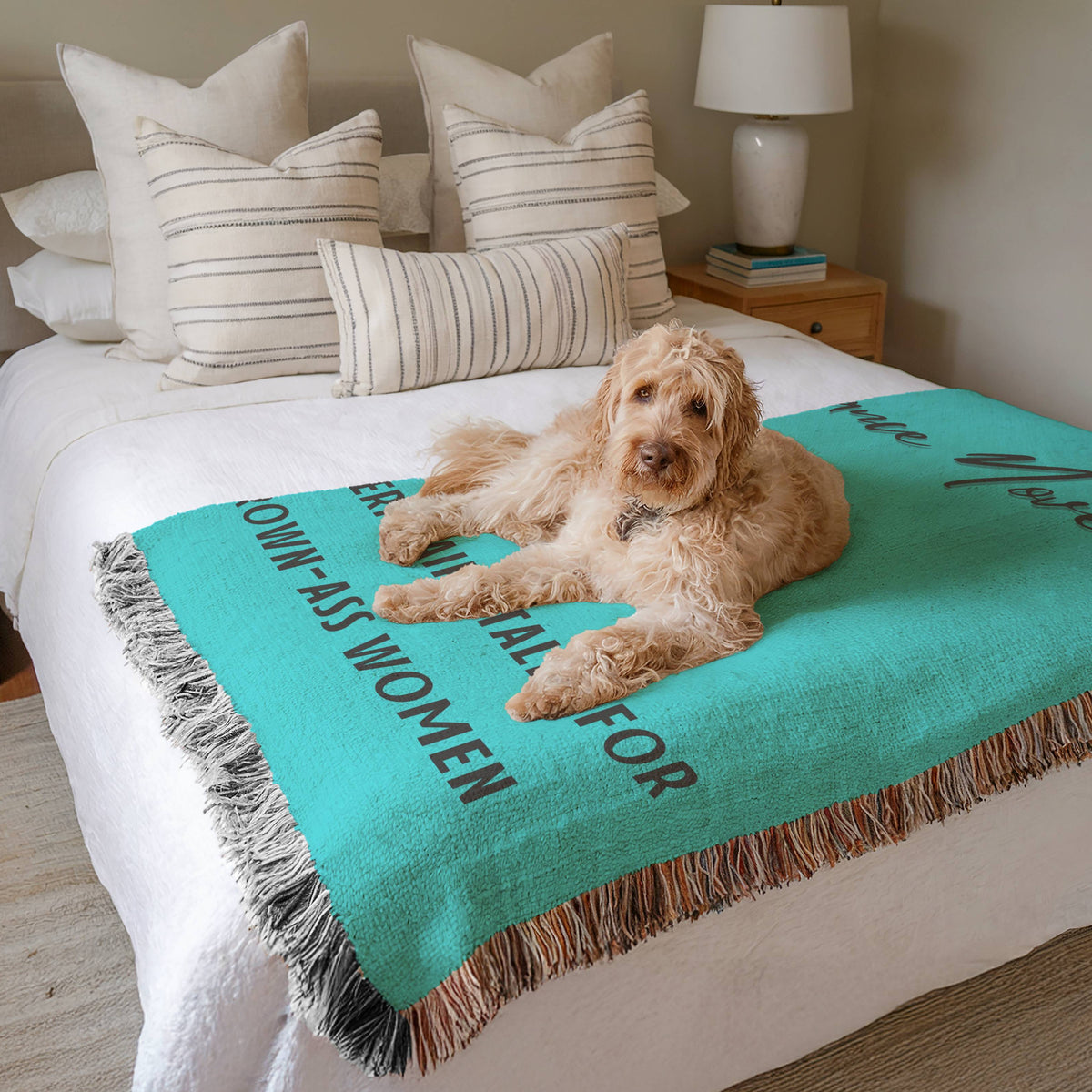 The blanket is displayed neatly folded on a bed with a white bedspread and striped pillows and a large white dog laying on it.