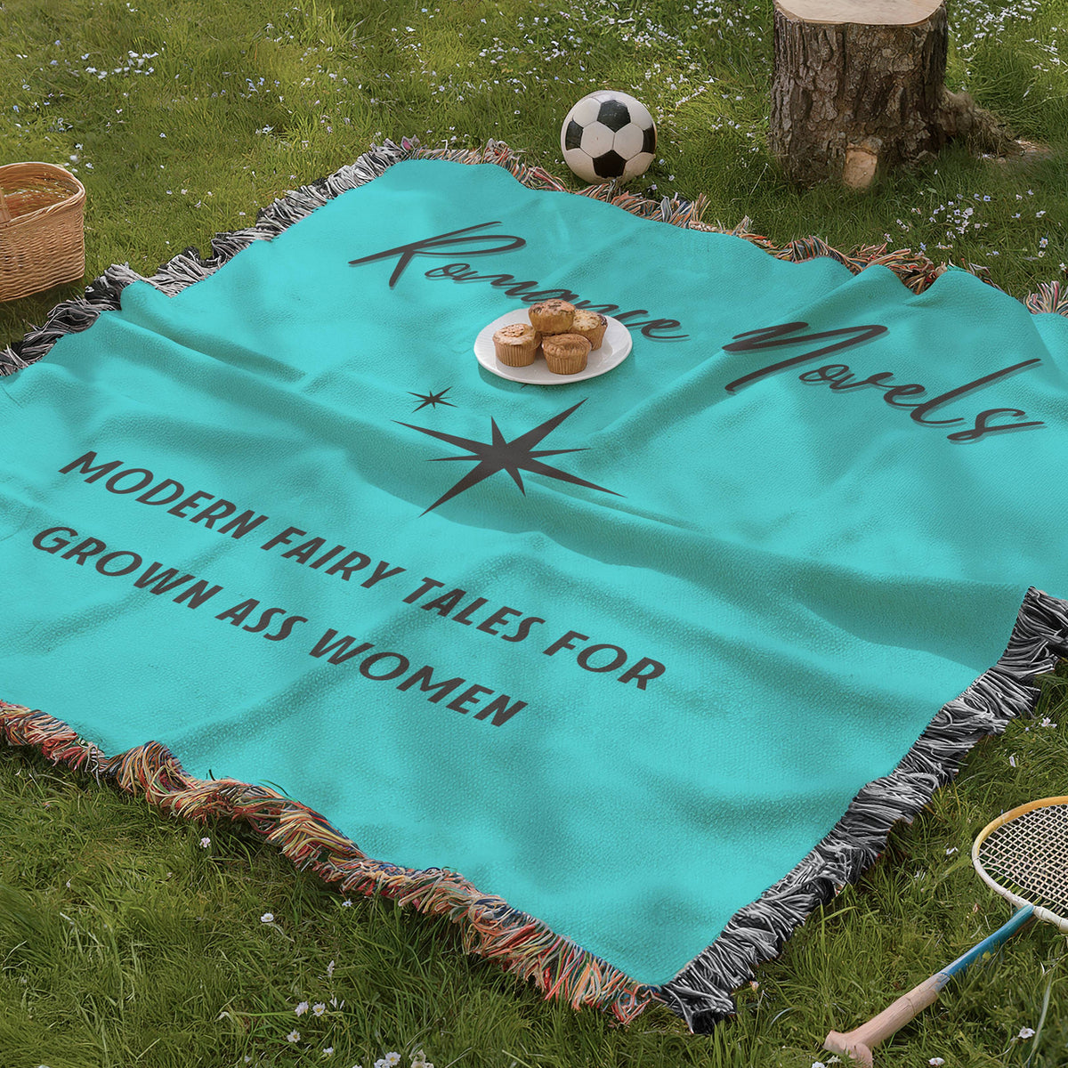 The blanket is being displayed as a picnic blanket in a park setting with a tree stump, picnic basket, plate of muffins, and a tennis racquet and a soccer ball lying around.
