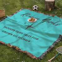 The blanket is being displayed as a picnic blanket in a park setting with a tree stump, picnic basket, plate of muffins, and a tennis racquet and a soccer ball lying around.
