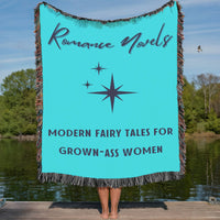 This turquoise-blue cozy woven blanket features the phrase “Romance Novels” in gray vintage typography above a centered whimsical retro starburst accent. Underneath the starburst in a bold, plain gray text are the words: “Modern Fairy Tales For Grown-Ass Women”. The blanket also features a colorful cotton fringe on all sides. It is being displayed by a woman who is showing it outstretched behind her. She is standing on a lakefront dock in a lake setting. The only visible part of her are her legs.   