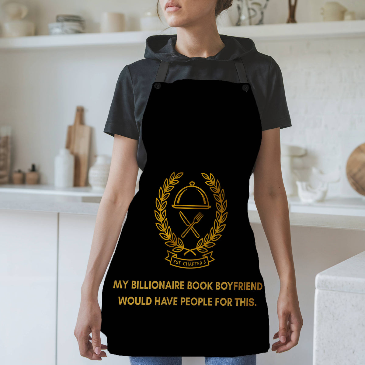 A woman wearing a black kitchen apron with a gold food service wreath graphic and the text 'MY BILLIONAIRE BOOK BOYFRIEND WOULD HAVE PEOPLE FOR THIS.' in a bright, modern kitchen setting.
