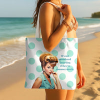 A woman on a beach holds a white tote bag with large mint green polka dots and a vintage-style illustrated woman on the front, next to the text, 'It's not considered hoarding if they're romance novels.'.