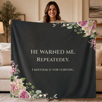 A woman stands in a living room holding up a dark charcoal sweatshirt blanket with light grey text that reads 'HE WARNED ME. REPEATEDLY. Then in smaller font, "I Mistook It For Flirting".  There is a small floral motif in the lower left and upper right corner.