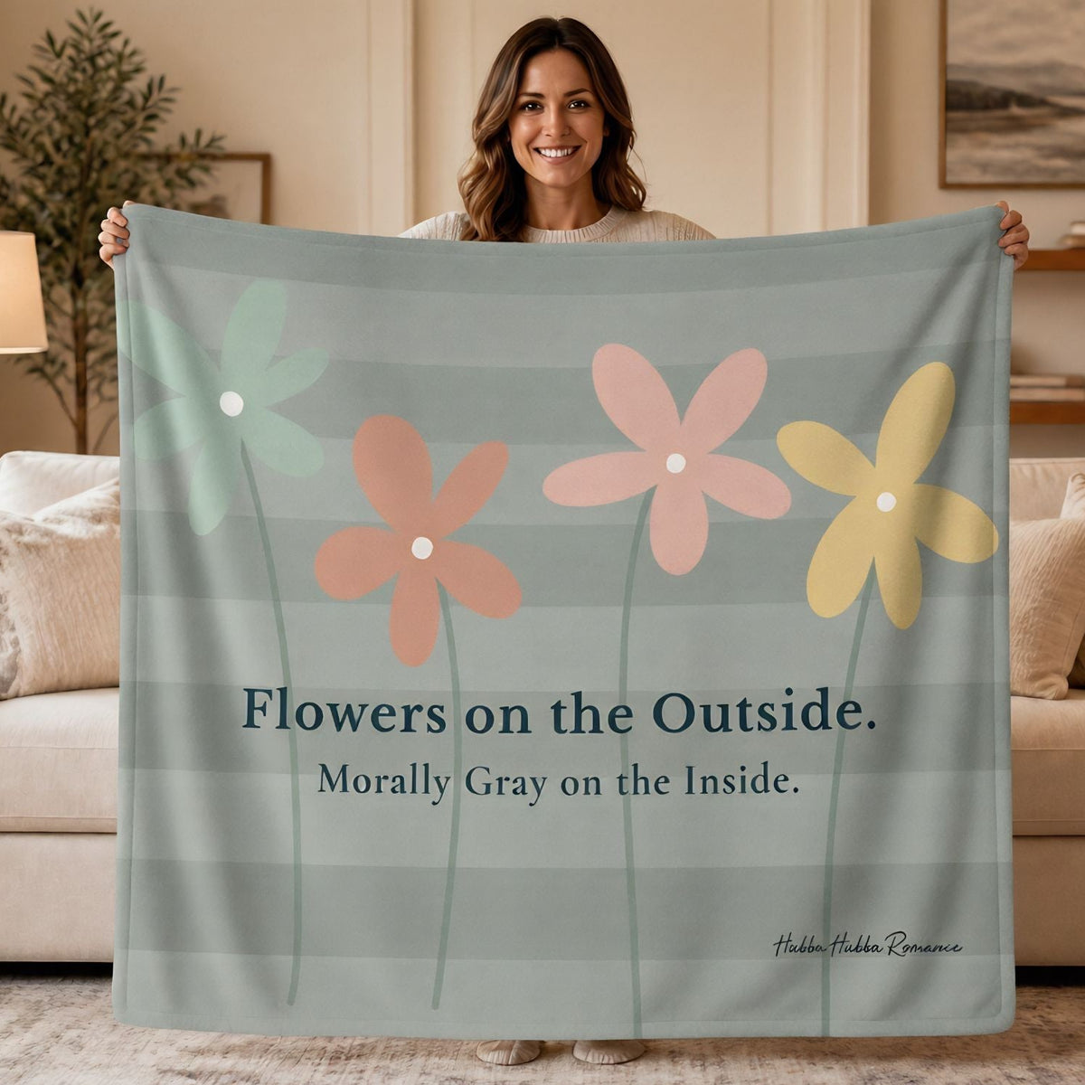 A woman stands in a living room holding up a large sweatshirt blanket. The blanket has a muted sage green  and light gray striped background with four stylized flowers in light saget green, coral pink, peach, and yellow, along with the text 'Flowers on the Outside. Morally Gray On The Inside.