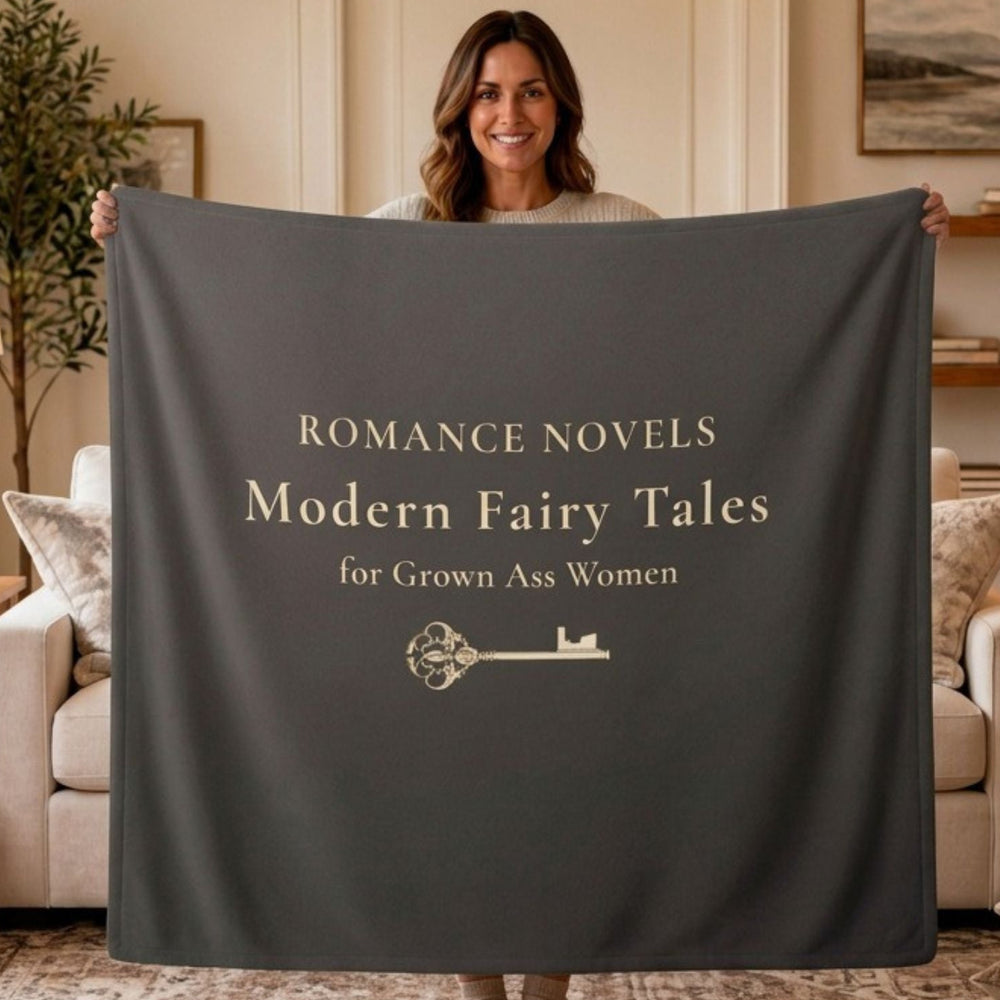 A woman stands in a living room holding up a large dark gray blanket or throw that reads, 'ROMANCE NOVELS Modern Fairy Tales for Grown Ass Women' with a small key graphic below.