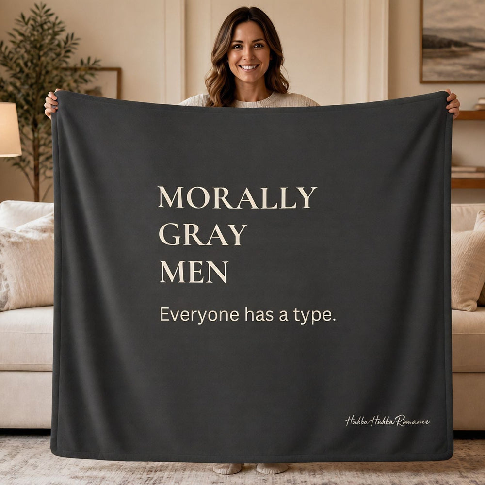 A woman stands in a living room holding up a large dark charcoal throw blanket with cream text that reads 'MORALLY GRAY MEN Everyone has a type.' A small script signature 'Abdel Halik Ramzan' is visible in the lower right corner of the blanket.