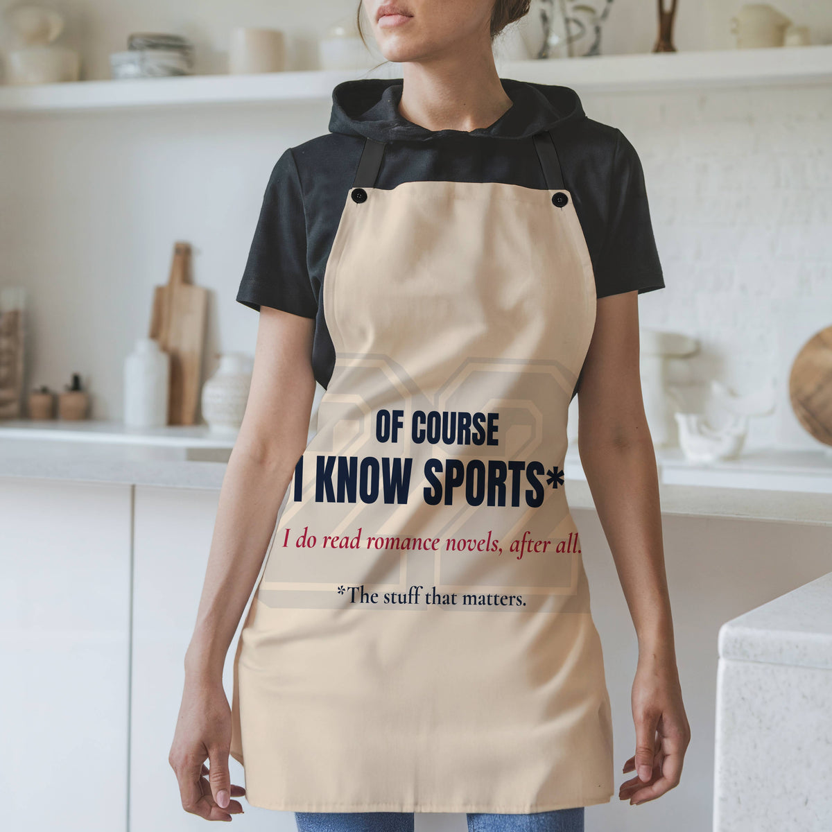 A woman wearing a beige apron over a black t-shirt in a bright kitchen setting. The apron features the text 'OF COURSE I KNOW SPORTS*' in dark blue, with red  text below reading 'I do read romance novels, after all. The last line, in dark blue, reads*the stuff that matters. There is a very faint 22 in a sports font behind the text.