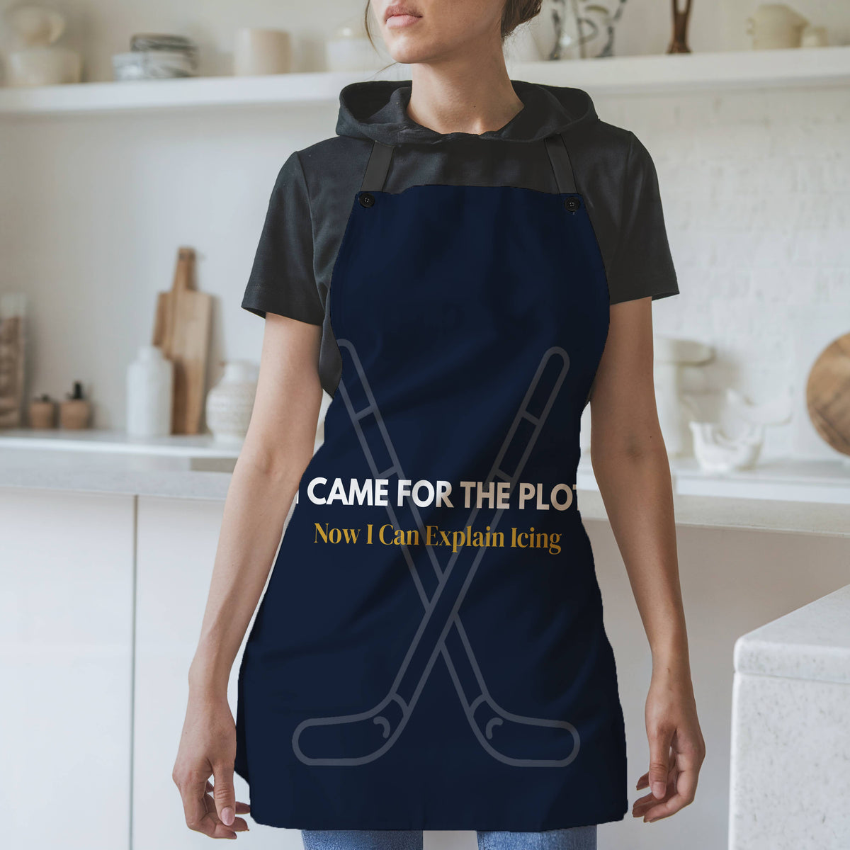 A woman wearing a navy blue apron with a humorous hockey-themed slogan, standing in a bright, modern kitchen. The dark navy blue apron with black neck and waist ties, features a graphic of crossed hockey sticks and the text 'I CAME FOR THE PLOT in white text. Now I Can Explain Icing' in gold.
