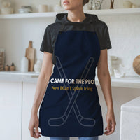 A woman wearing a navy blue apron with a humorous hockey-themed slogan, standing in a bright, modern kitchen. The dark navy blue apron with black neck and waist ties, features a graphic of crossed hockey sticks and the text 'I CAME FOR THE PLOT in white text. Now I Can Explain Icing' in gold.