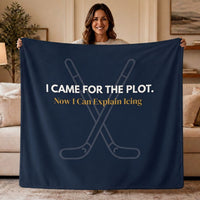 A woman holds up a navy blue blanket in a living room setting. The blanket features white text that reads 'I CAME FOR THE PLOT. Underneath it gold text reads "Now I can explain icing."