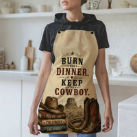 A woman wearing a beige apron with a western-themed graphic print that reads 'BURN THE DINNER. KEEP THE COWBOY.