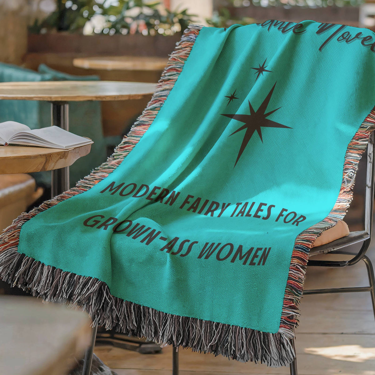 The blanket is displayed lying on a chair in a coffee shop setting with an open book and a cup of coffee on a small round table.

