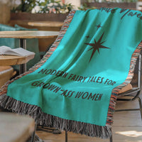 The blanket is displayed lying on a chair in a coffee shop setting with an open book and a cup of coffee on a small round table.

