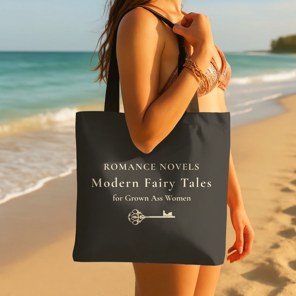 A woman on a beach holds a dark gray tote bag with cream text that reads 'ROMANCE NOVELS Modern Fairy Tales for Grown Ass Women' and a royal small key graphic under the text.