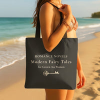 A woman on a beach holds a dark gray tote bag with cream text that reads 'ROMANCE NOVELS Modern Fairy Tales for Grown Ass Women' and a royal small key graphic under the text.
