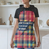 A woman wearing a black t-shirt and a colorful plaid apron with the text 'NOW SERVING SMUT. to be followed BY DINNER.