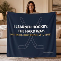 A woman stands in a living room holding a navy blue sweatshirt  blanket that reads, 'I LEARNED HOCKEY. THE HARD WAY. One book boyfriend at a time.