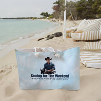The weekender bag is displayed standing upright on the beach next to beach towels.
