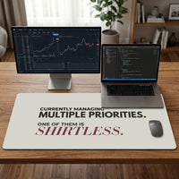 A large taupe desk mat with humorous text sits on a wooden desk, styled with a free standing computer monitor, a laptop, and wireless mouse. The desk mat features the text 'CURRENTLY MANAGING MULTIPLE PRIORITIES. One of them is Shirtless.