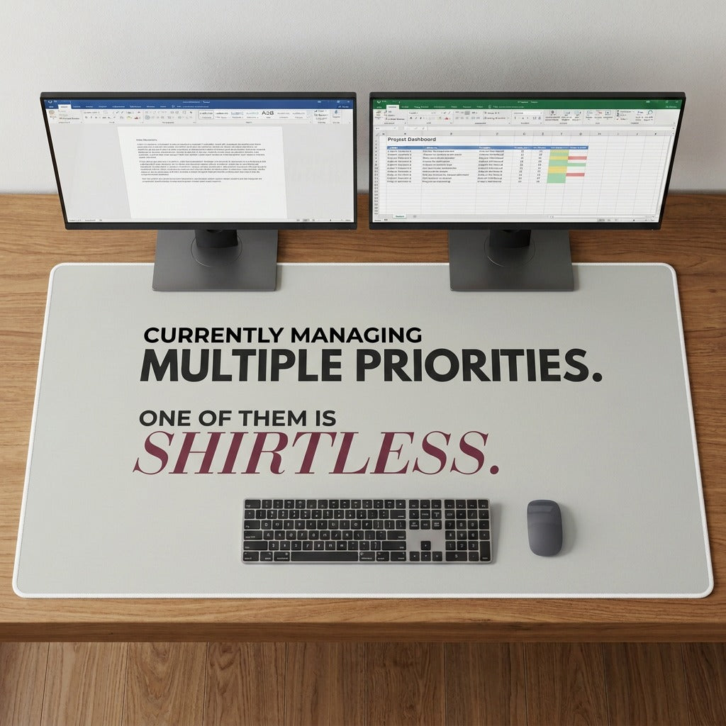An oversized desk mat with a humorous quote printed on it, displayed on a wooden desk with two full size free standing computer monitors, a keyboard, and a mouse. The text says Currently Managing Multiple Priorites. One of them is shirtless.