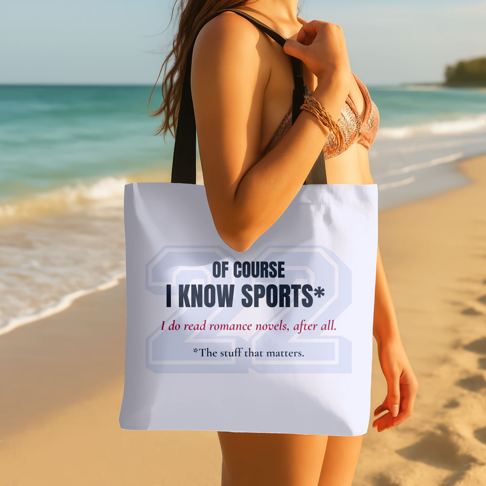 Pale blue romance reader tote bag with a large pale gray varsity-style 22 and dark navy text reading “Of Course I Know Sports*.” Red script reads “I do read romance novels, after all.” Small dark text reads “*The stuff that matters.” Black handles. Carried by a woman on a sandy beach with the ocean in the background.
