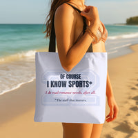 Pale blue romance reader tote bag with a large pale gray varsity-style 22 and dark navy text reading “Of Course I Know Sports*.” Red script reads “I do read romance novels, after all.” Small dark text reads “*The stuff that matters.” Black handles. Carried by a woman on a sandy beach with the ocean in the background.