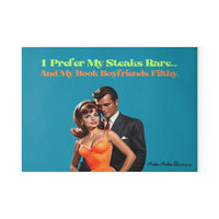 Glass cutting board with a turquoise blue background. Features a glamorous vintage couple, the woman in a orange dress and the man in a dark suit. The text above them reads: “I prefer my steaks rare” in a lime green vintage text... “and my book boyfriends filthy in a bright orange vintage text.” Made of durable tempered glass with non-slip rubber feet for countertop use. This is the smaller size.