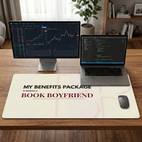 A large cream-colored desk mat with the text 'MY BENEFITS PACKAGE IS MISSING A BOOK BOYFRIEND' in black and burgundy lettering, shown on a wooden desk with a laptop and second free standing monitor to the left.