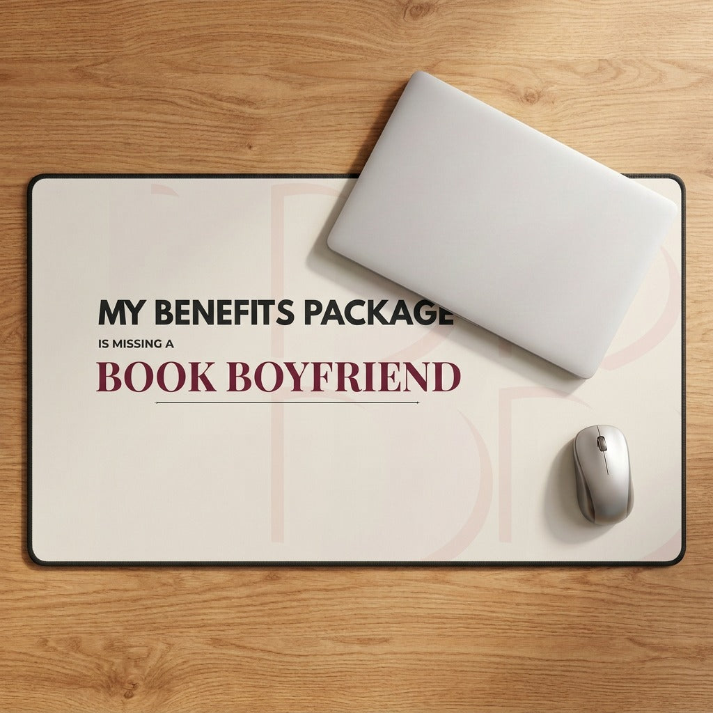 A rectangular desk mat with a humorous book-themed message reading 'MY BENEFITS PACKAGE IS MISSING A BOOK BOYFRIEND' in black and burgundy text, styled on a wooden desk with a closed silver laptop and a white computer mouse.