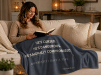 Woman reading on a cozy sofa while wrapped in a dark romance sweatshirt blanket displaying the quote "He's Cursed. He's Dangerous. He's Morally Compromised." Comfortable reading blanket for fans of morally gray men and dark romance books.