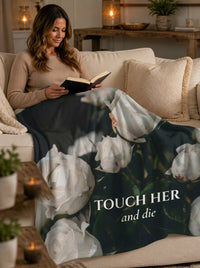 A woman sits on a beige sofa reading a book while wrapped in a dark floral blanket with large white rose imagery and the text 'TOUCH HER and die'.