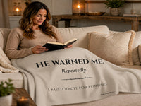Romance reader relaxing on a sofa while wrapped in a cream-colored sweatshirt blanket featuring the quote "He Warned Me. Repeatedly. I Mistook It For Flirting." Cozy reading scene inspired by dark romance books, morally gray men, and fictional red flags.