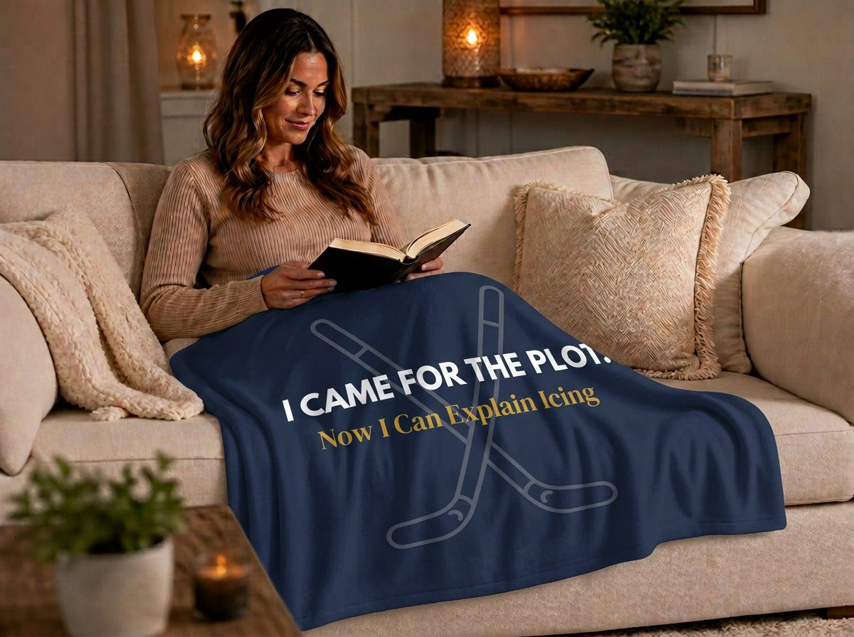 A woman sits on a beige sofa reading a book while wrapped in a navy blue blanket with white and yellow text that reads, 'I CAME FOR THE PLOT. Now I Can Explain Icing' in front of faded crossed hockey sticks.