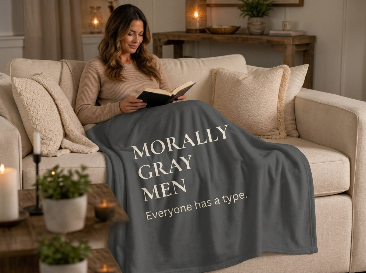 A woman sits on a beige sofa in a cozy living room, wrapped in a dark gray sweatshirt  blanket printed with light grey text 'MORALLY GRAY MEN Everyone has a type.'. 
