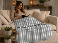 A woman sits on a beige sofa in a cozy living room, reading a book while wrapped in a gray and white striped blanket printed with the text 'MORALLY GRAY MEN are just my type. So call me crazy.