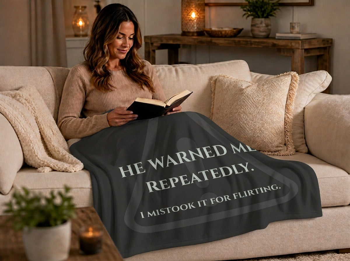 A woman sits on a beige sofa reading a book, wrapped in a dark charcoal throw blanket printed with light gray text that reads, 'HE WARNED ME REPEATEDLY. I mistook it for flirting. There is a faded  gray warning-triangle graphic behind the text.