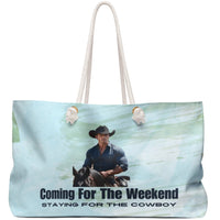 Image shows a muscular cowboy in a dark blue button-down and black hat riding a black horse against a soft watercolor sky blue background. Below the image is bold black text reading: “Coming for the Weekend,” followed by smaller, italicized text: “Staying for the Cowboy.” The design evokes a western romance aesthetic, blending humor and heat. Displayed on a weekender bag, it targets romance readers who adore cowboy tropes, rugged heroes, and a little weekend mischief.