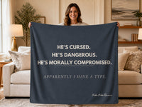 Woman standing in a living room holding a dark blue sweatshirt blanket with the quote "He's Cursed. He's Dangerous. He's Morally Compromised. Apparently I Have a Type." Blanket designed for dark romance readers and fans of morally gray heroes.
