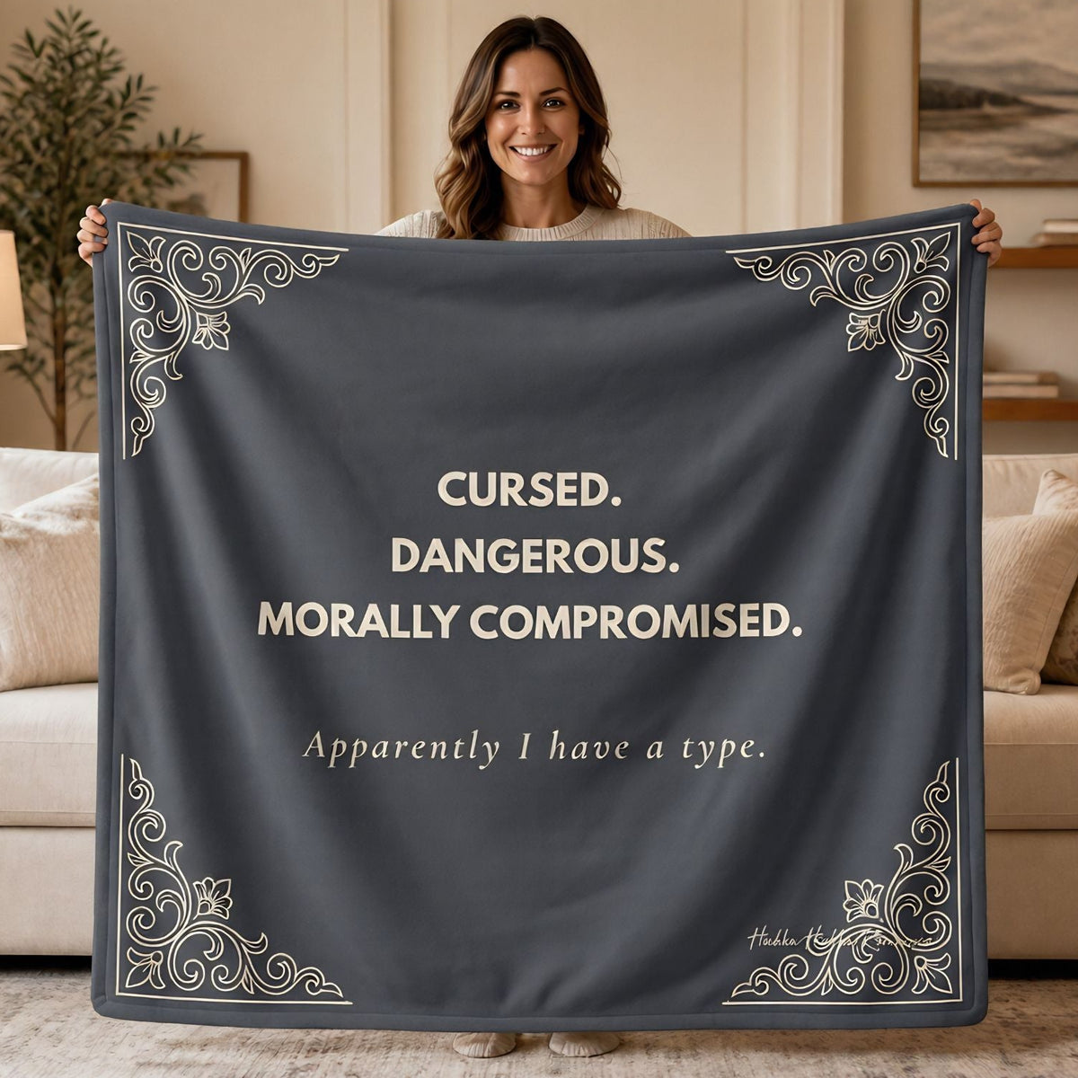 Woman holding a slate gray sweatshirt blanket with the words "Cursed. Dangerous. Morally Compromised. Apparently I have a type." in a cream colored font and ornate gothic corner details for dark romance readers.