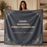Woman holding a slate gray sweatshirt blanket with the words "Cursed. Dangerous. Morally Compromised. Apparently I have a type." in a cream colored font and ornate gothic corner details for dark romance readers.