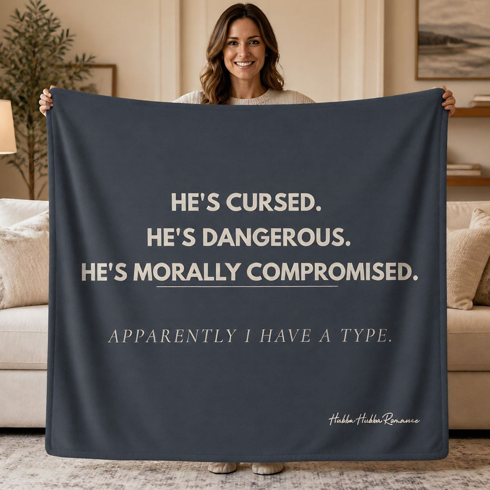 A woman stands in a living room holding up a dark navy blanket or throw with cream text that reads, 'HE'S CURSED. HE'S DANGEROUS.Apparently I have A Type
