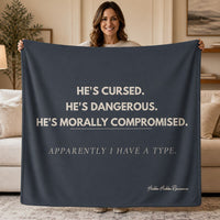 A woman stands in a living room holding up a dark navy blanket or throw with cream text that reads, 'HE'S CURSED. HE'S DANGEROUS.Apparently I have A Type
