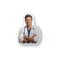 ProducCustom-shaped decorative pillow featuring The Doctor, a handsome medical romance book boyfriend with salt-and-pepper hair, light stubble, and a confident smile, wearing a white lab coat with a stethoscope and folded arms. Shown as a standalone pillow against a neutral background. This medical romance book boyfriend pillow is available in three sizes (10" x 10", 16" x 16", and 22" x 22").t mockup