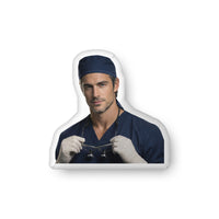 Custom-shaped decorative pillow featuring The Sexy Surgeon, a medical romance book boyfriend with dark hair, light stubble, and a confident expression, wearing navy surgical scrubs and gloves while holding surgical loupes. Shown as a standalone pillow against a neutral background. This medical romance book boyfriend pillow is available in three sizes (10" x 10", 16" x 16", and 22" x 22").