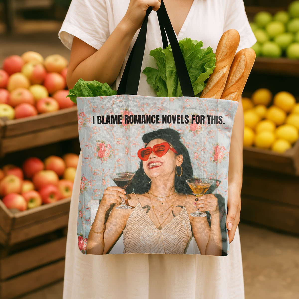 A person in a white dress holds a tote bag in a grocery store produce section. The tote bag has a light blue floral background with a printed image of a woman in a beaded dress holding a cocktail in each hand, with text above that says "I BLAME ROMANCE NOVELS FOR THIS.