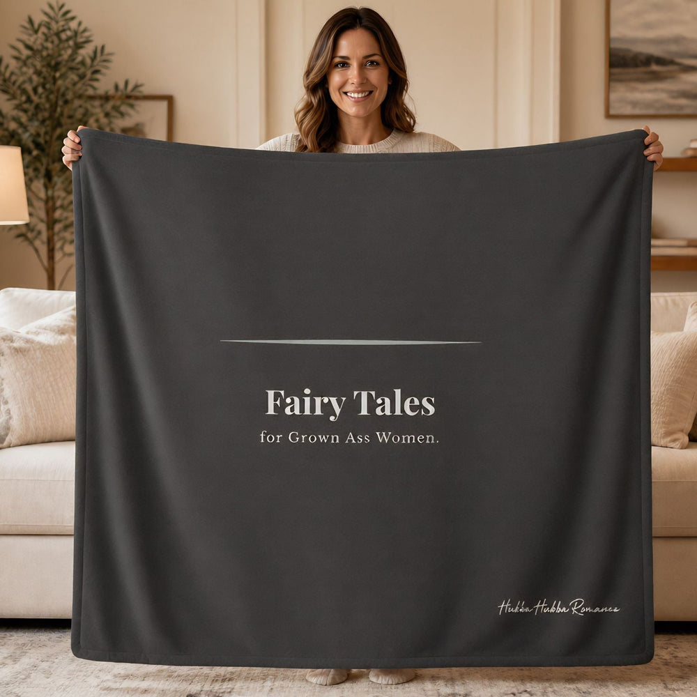A woman stands in a living room holding up a large dark charcoal gray sweatshirt blanket. The blanket features the text 'Fairy Tales for Grown Ass Women.