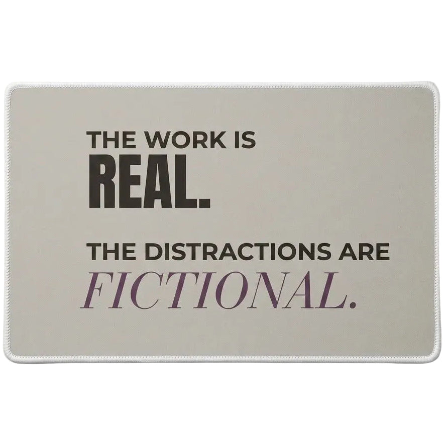 A rectangular desk mat with a taupe  background and a white stitched border. The mat features the text 'THE WORK IS REAL.The Distractions Are Fictional.