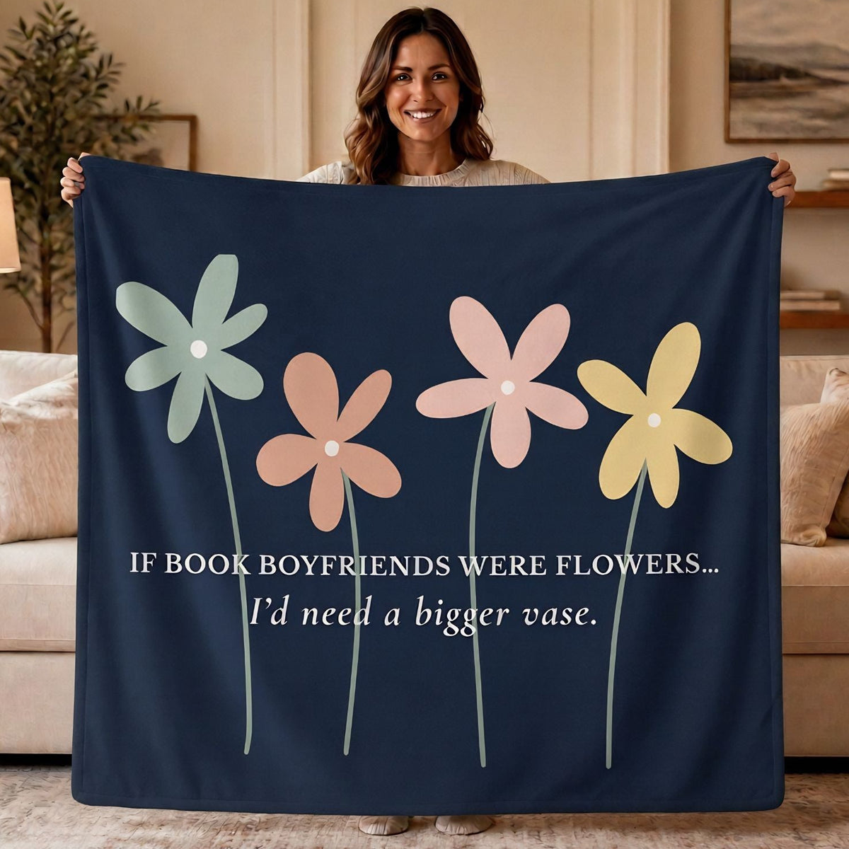 A woman holds up a navy blue blanket in a cozy living room setting. The blanket features four colorful pastel flowers (light blue, peach, pink, and yellow) and white text that reads, 'IF BOOK BOYFRIENDS WERE FLOWERS. I'd need a bigger vase